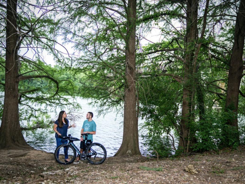 A student and a professor stand by Lady Bird Lake with a bike and are surrounded by trees.