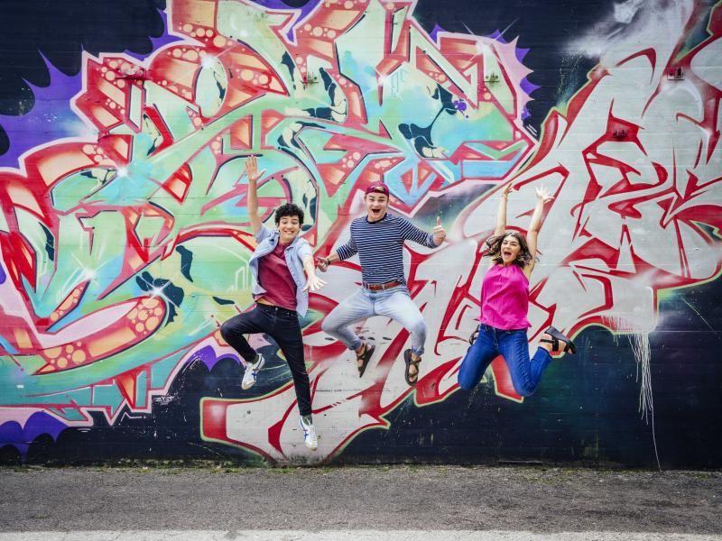 Three students jump in front of a colorful, patterned mural.