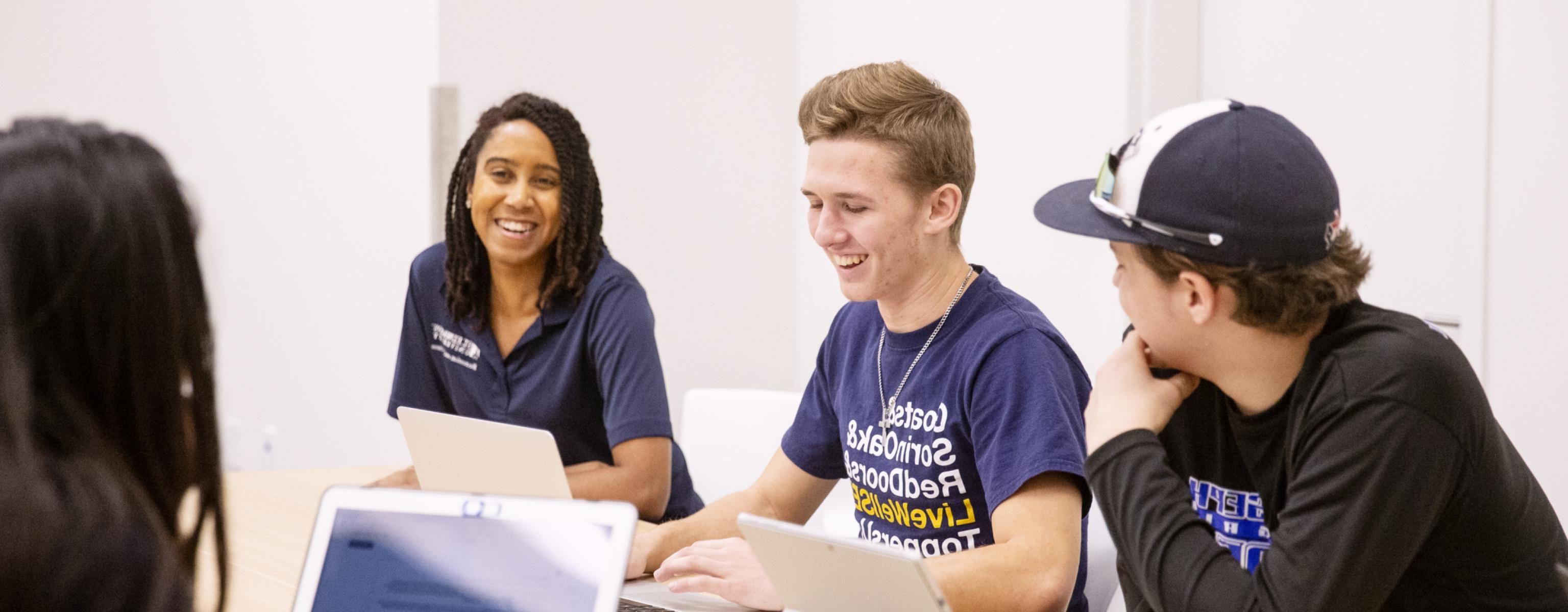 Students and a staff member smile during a meeting as they sit at a meeting table with laptops and tablets.