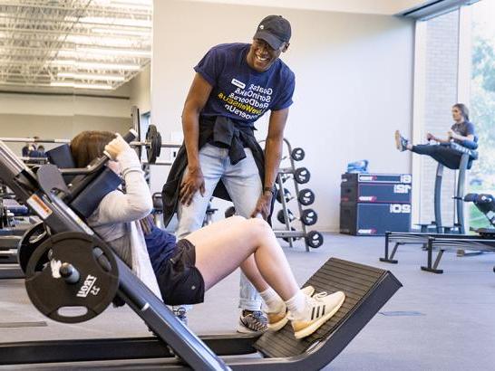 A peer training coach stands gives a student instructions on a hack squat press. Another student is in the background performing hanging leg raises.