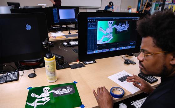 The image s如何s a man sitting at a desk in a computer lab, working on graphic design. He has short curly hair, glasses, 和 is focused on his work. 在他的桌子上有两个电脑显示器, 显示骨架图形的一个, 另一组是空白屏幕. He has a green stencil with a skeleton design taped to the desk, 还有一些工具，比如卷尺和罐子. Other workstations 和 people are visible in the background.